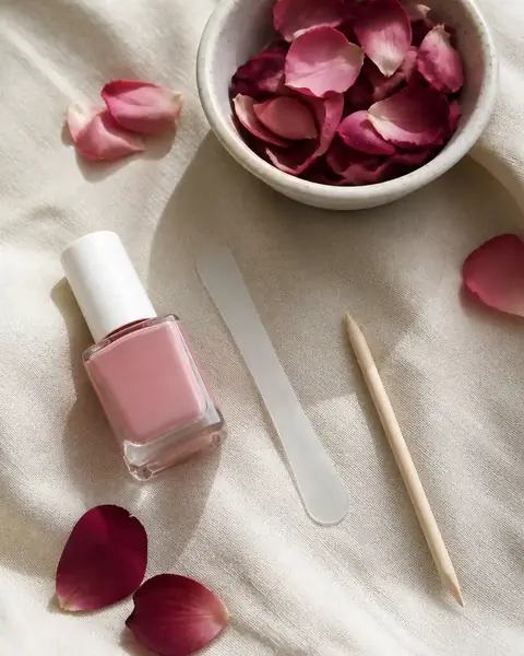Lima di vetro, smalto rosa nude, bastoncino d’arancio e una ciotola di petali su un panno di lino